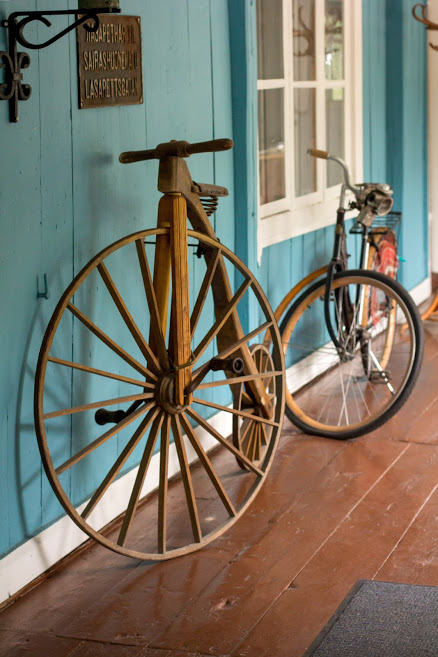 Museorakennuksen lasiveranta. Seinään nojaa vanha puinen pyörä, jossa on iso etupyörä ja pieni takapyörä.