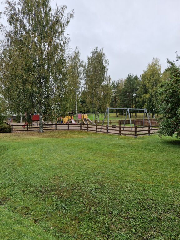 Kiikalan leikkipuistosta kuva, kaukaa otettu. Kuvassa näkyy leikkipuiston aita ja aidan sisällä keinut, liukumäki, pieni karuselli ja kiipeilypaikka juna.